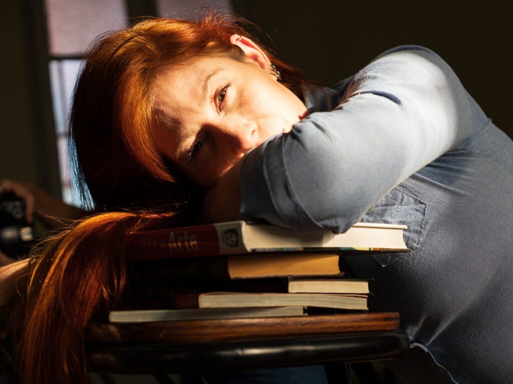 Woman resting her head on a stack of books, representing the exhaustion, low energy, and overwhelm that can occur with burnout and depression.